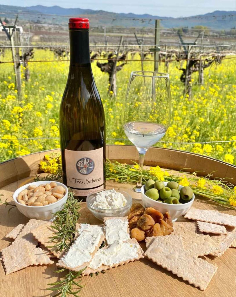 bottle of Tres Sabores Picpoul Blanc on a barrel with a glass and snacks